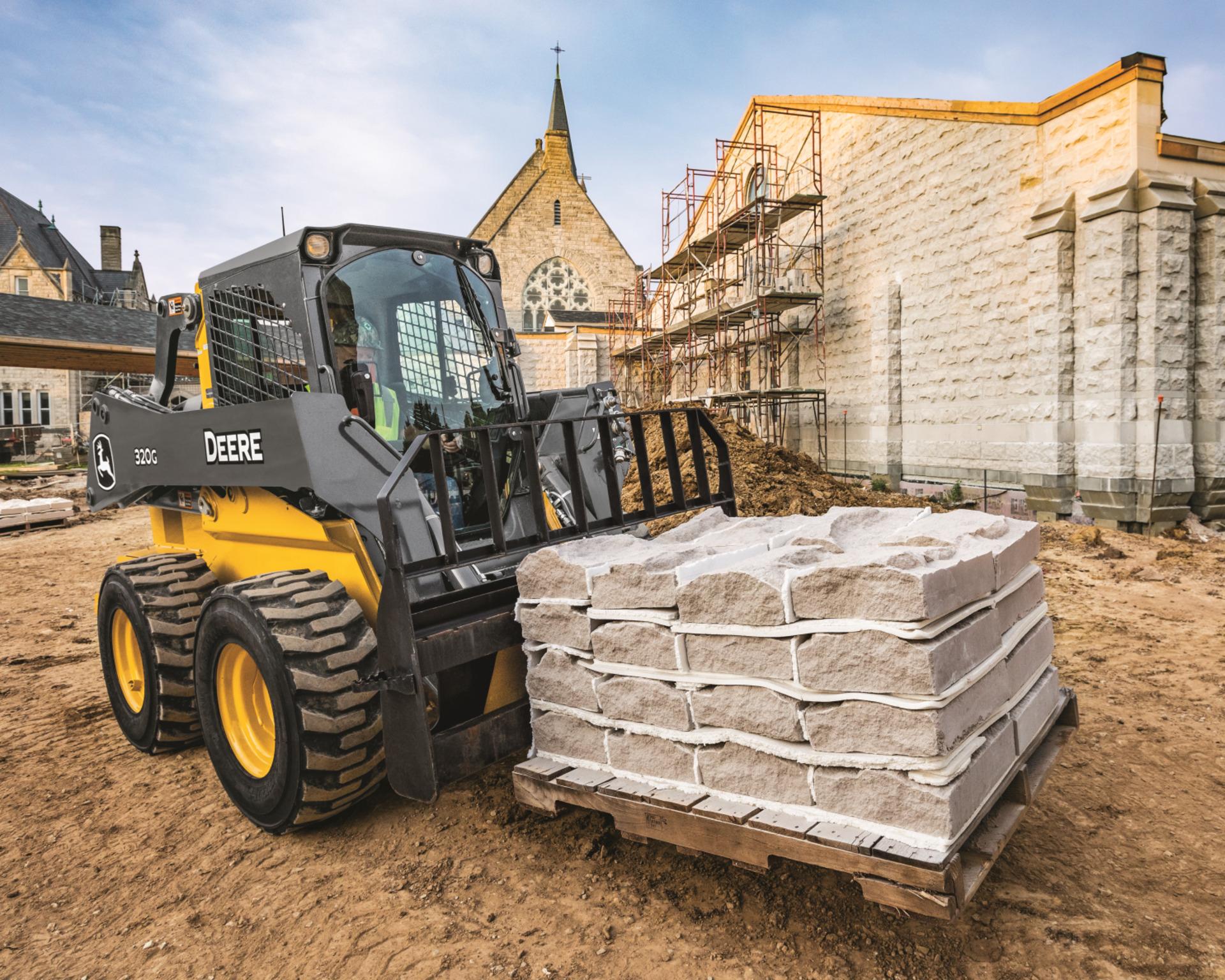 A John Deere 320G skid steer uses a pallet to move concrete blocks.