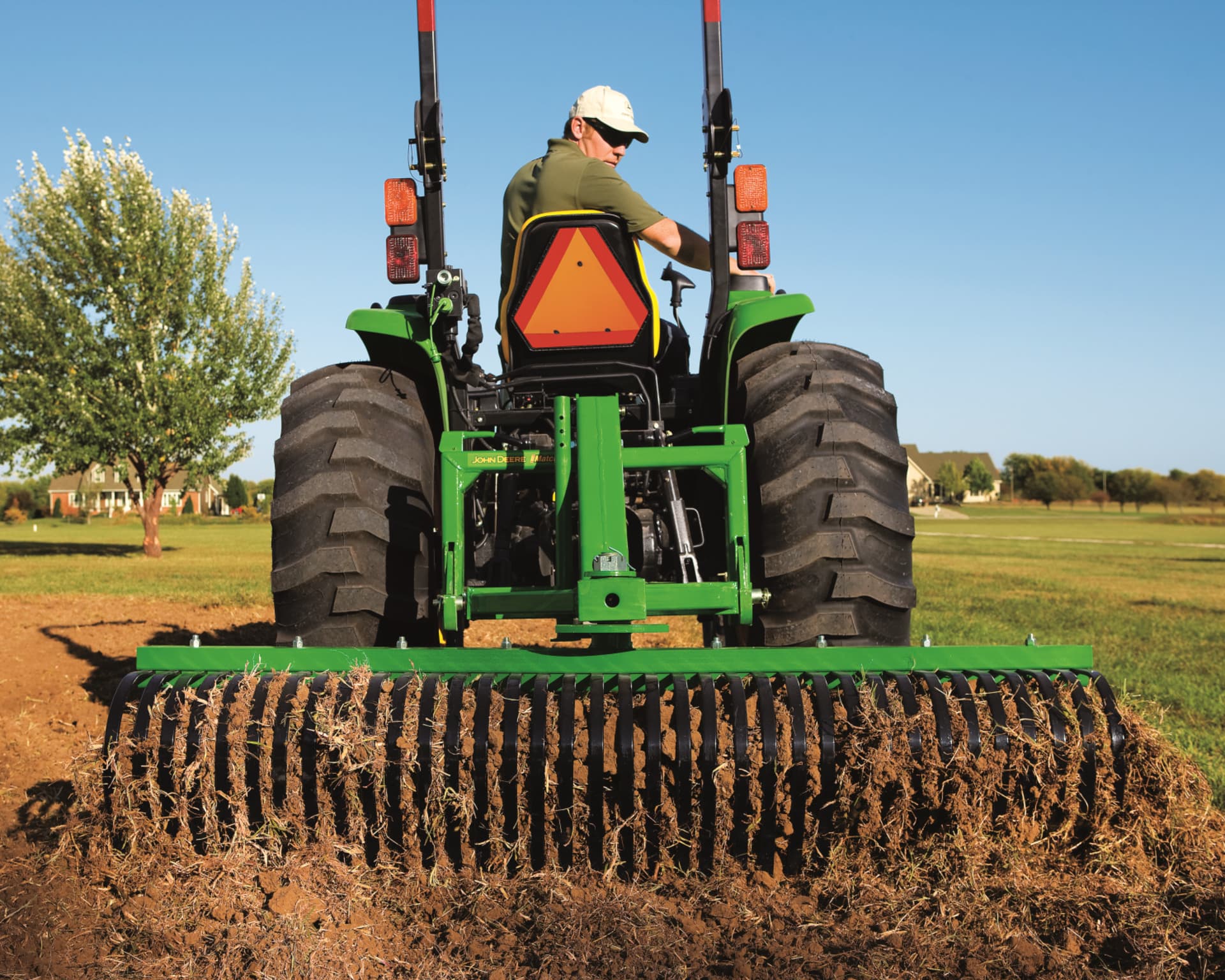 A homeowner uses a landscape rake attachment on a John Deere compact tractor.