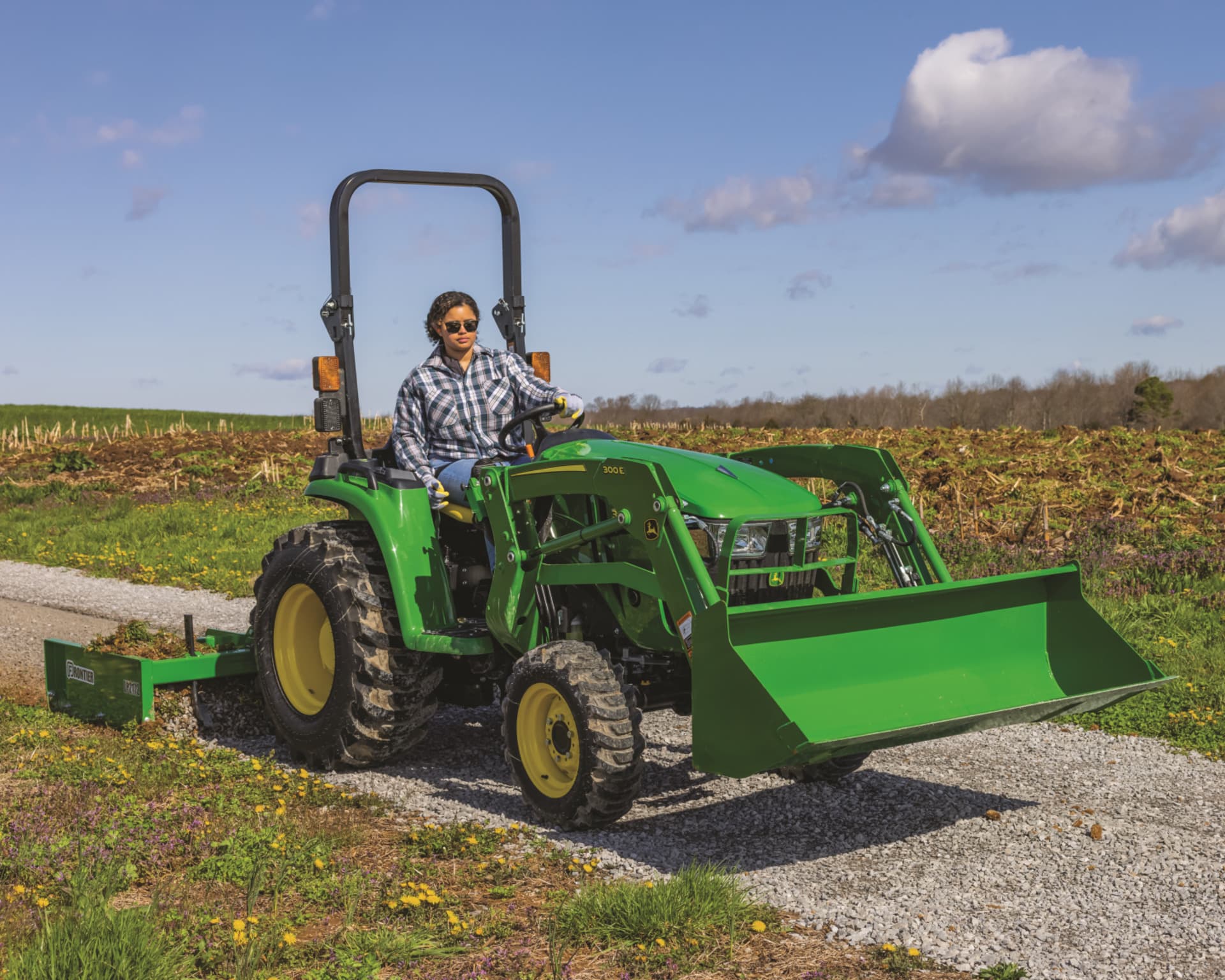 A person operates a John Deere compact tractor with a scraper and front-end loader attachments.
