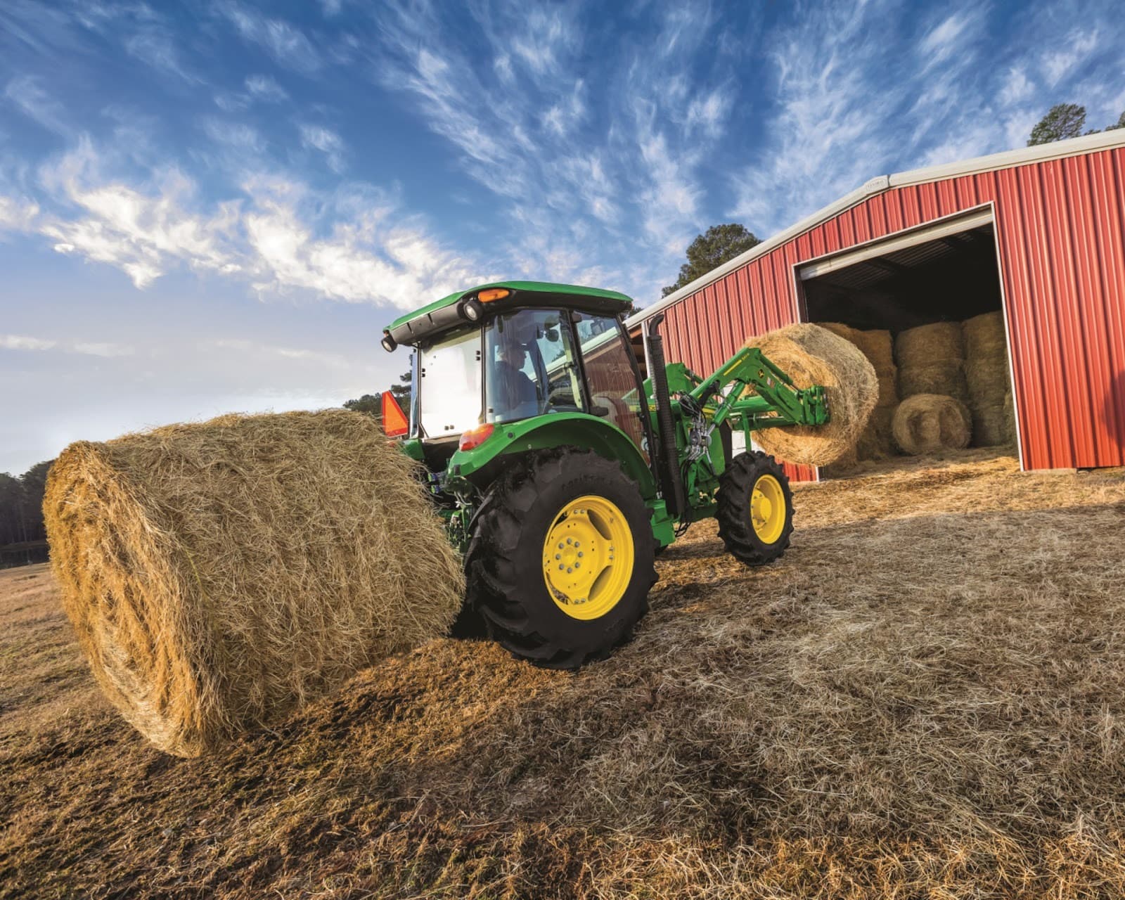 A John Deere tractor with a bale spear attachment moves a hay bale to a red barn.