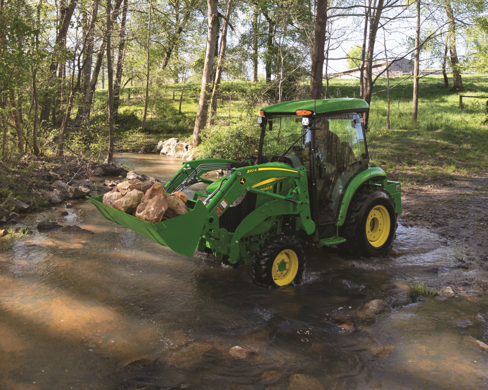 A John Deere compact utility tractor with a front-end loader lifts rocks on a property.