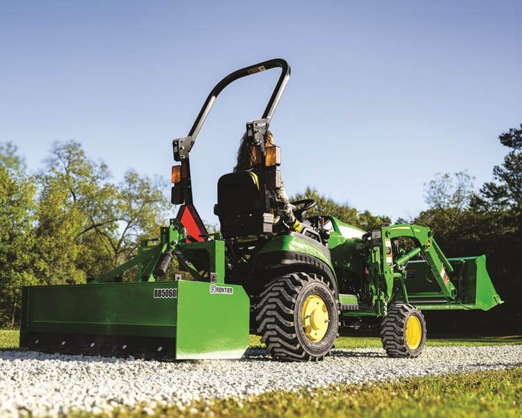 A property owner uses a box blade attachment on a John Deere tractor to grade their gravel driveway.