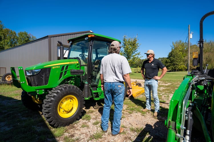 Two farm workers talk on the farm standing next to a John Deere 5 Series tractor.