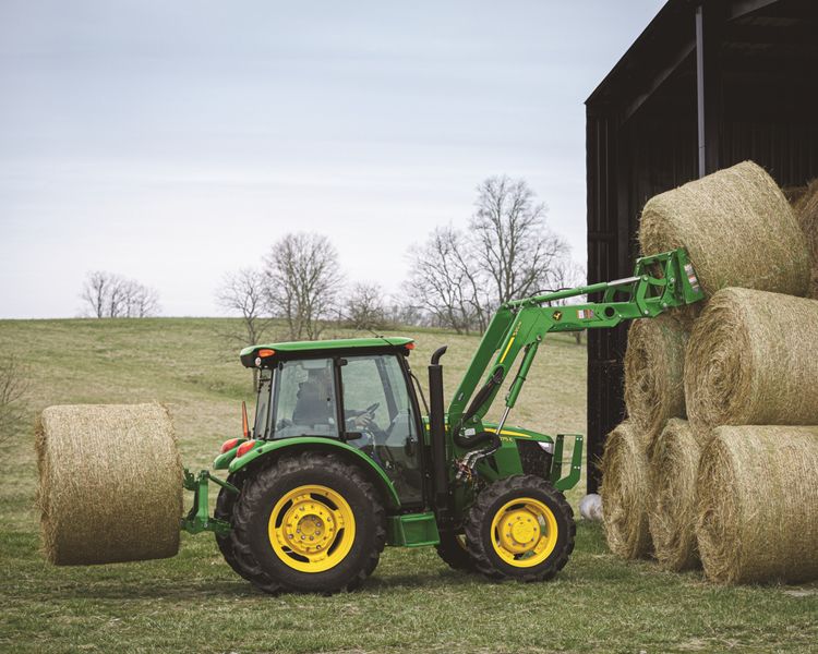 A John Deere 5075E lifting hay bails into a barn using a front end loader.