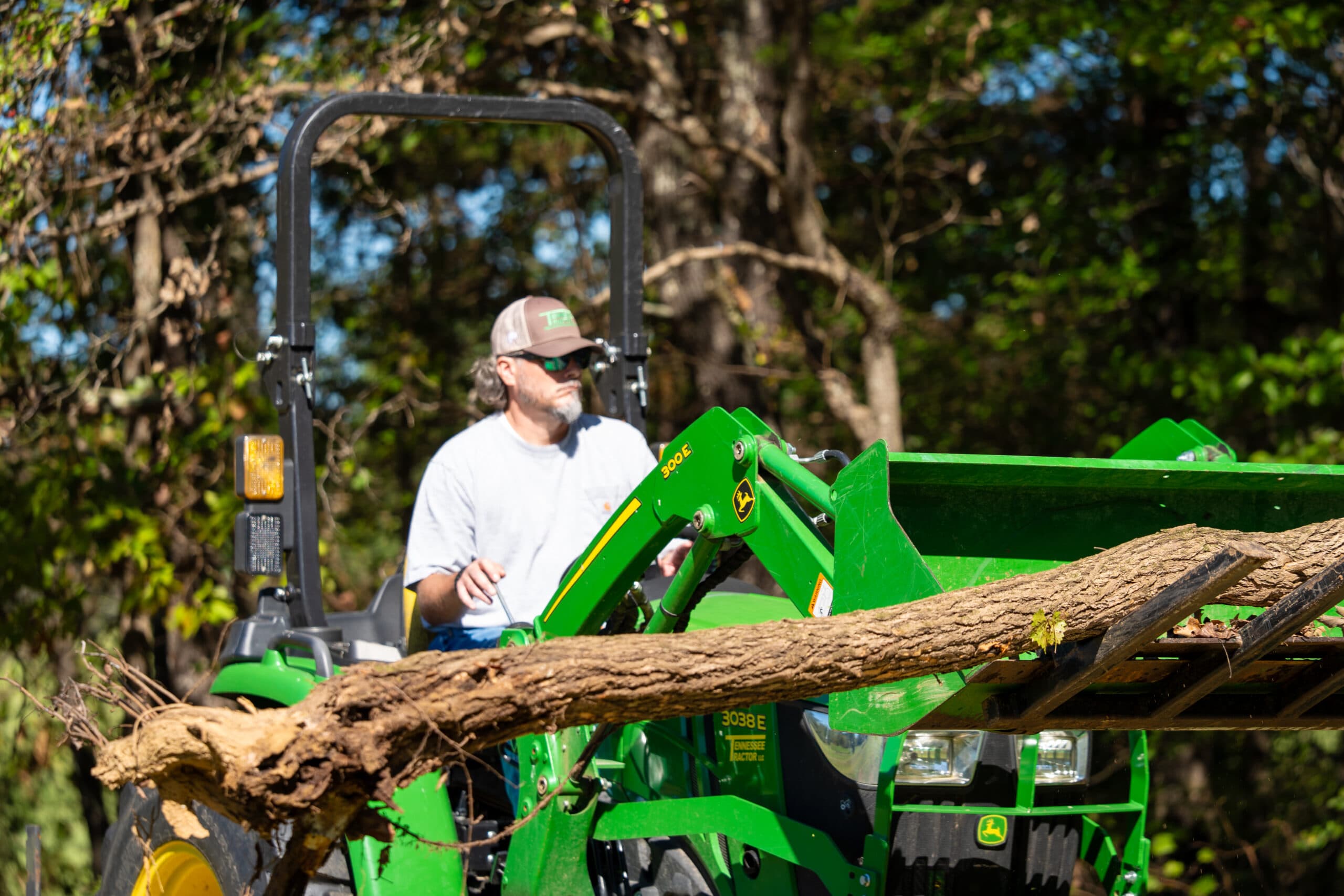 A man uses a John Deere tractor with a front-end loader to lift a tree branch.